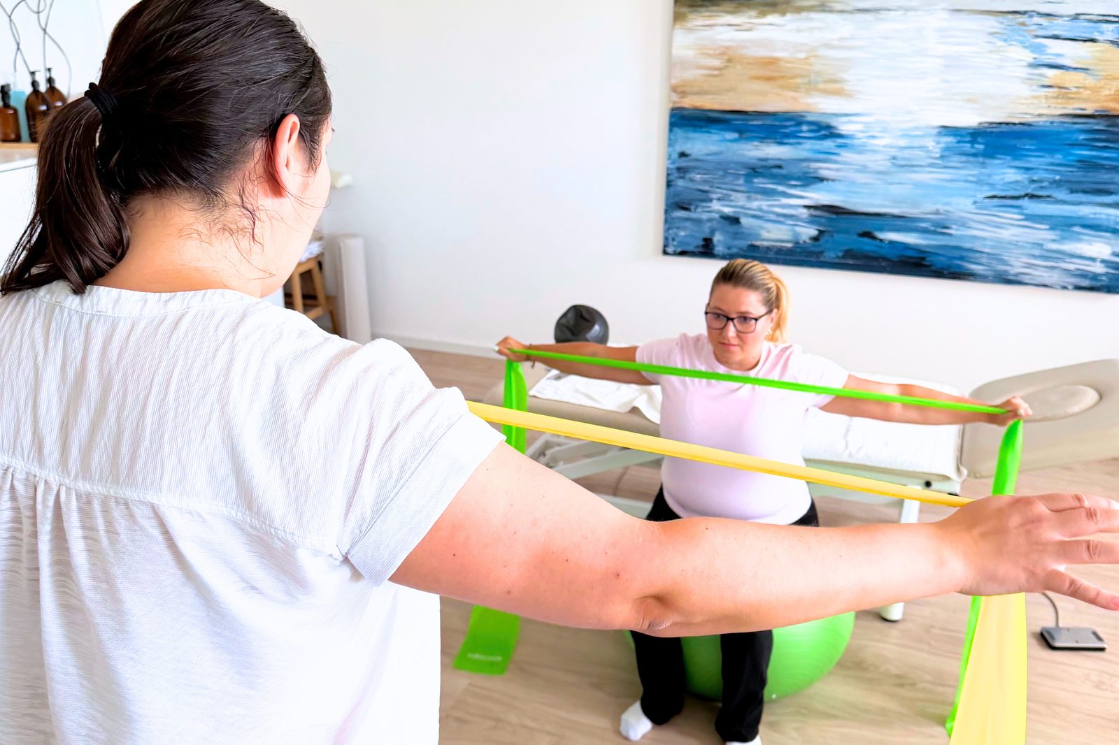 Lucie Barak zeigt eine Übung mit dem Theraband vor, die Patientin sitzt auf dem Gymnastikball.