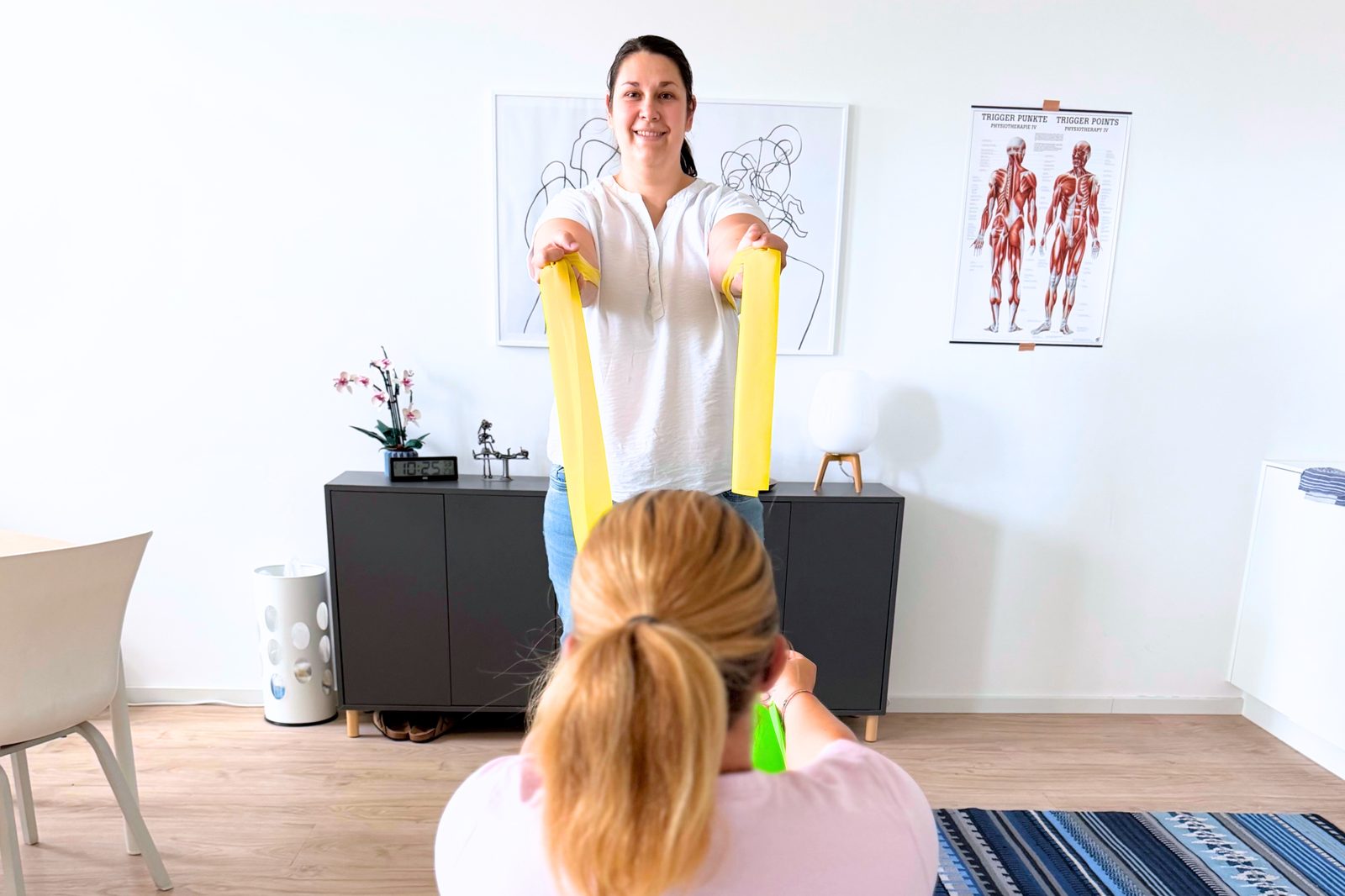 Eine Patientin trainiert mit dem Theraband, Lucie Barak führt die Übung vor.