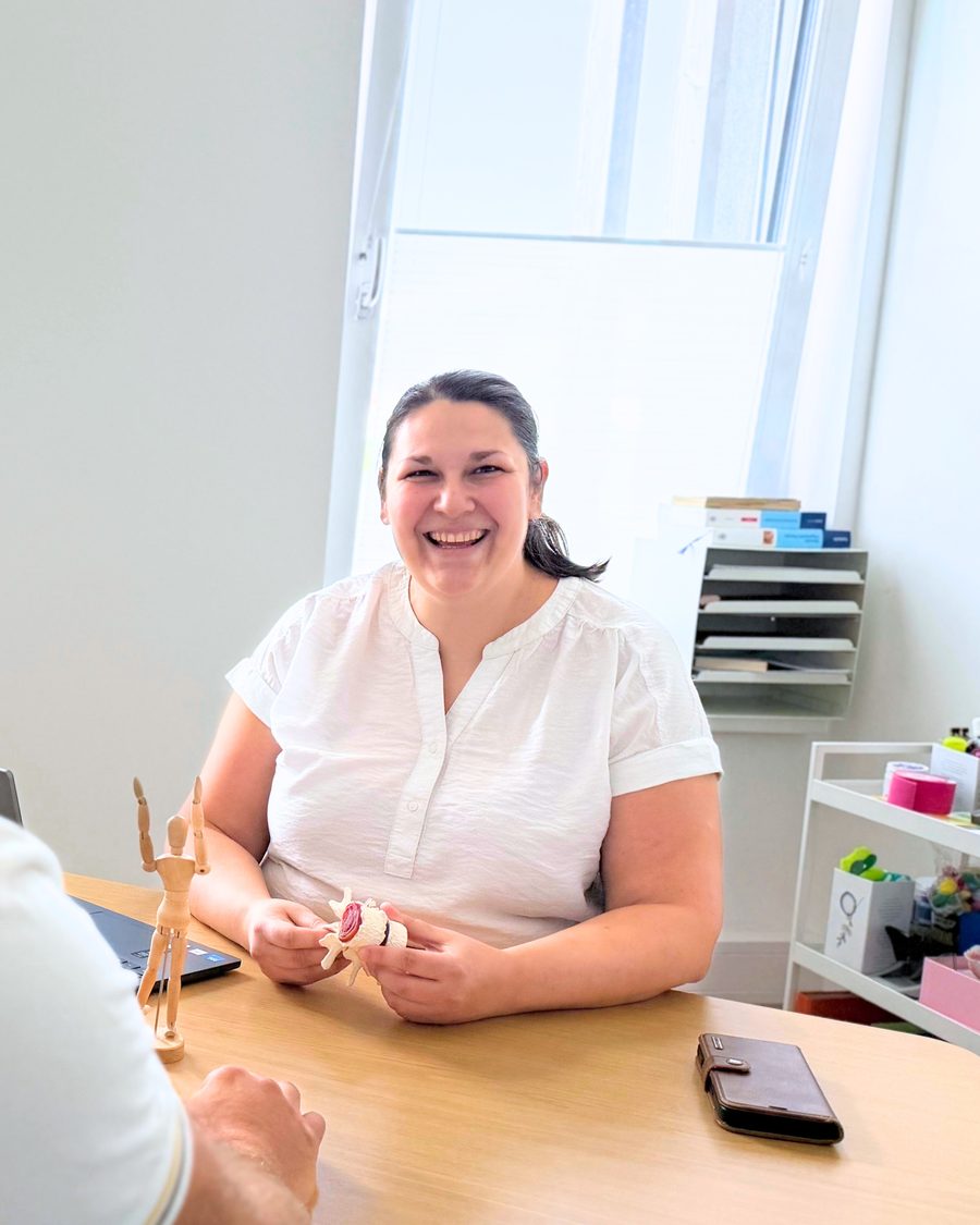 Lucie Barak im Gespräch mit einem Patienten am Schreibtisch, ein Wirbelsäulenmodell in der Hand.